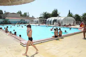 Außenpool auf einem Campingplatz mit Sonnenschirmen, Menschen im Wasser und einer überdachten Schwimmfläche im Hintergrund.