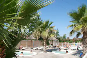 Camping-Pool mit Palmen im Vordergrund, Liegen und Sonnenschirmen am Beckenrand, mehrere Personen im Wasser.