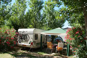 Wohnwagen auf einem Campingplatz mit Markise, zwei Stühlen und Tisch vor grünen Bäumen und Blumenbeeten.