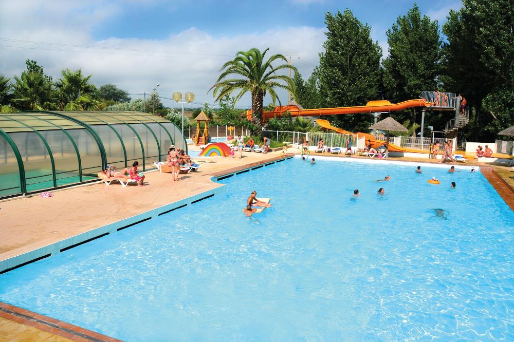 Großes Camping-Schwimmbecken mit orangefarbenem Wasserrutschenturm, Liegen und Palmen im Hintergrund.