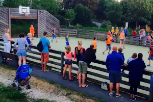 Kinder und Erwachsene auf einem Sportplatz am Campingplatz, mit Basketballkorb, Holzlattenzaun und Sitzbank am Rand.