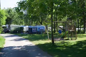 Campingplatz mit Wohnwagen und Zelt zwischen Bäumen, daneben Spielplatz mit Schaukel und Rutsche.