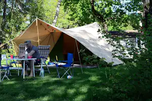 Campingzelt im Grünen mit Sonnensegel, Campingstühlen und Tisch am Stellplatz zwischen Bäumen und Hecken.