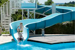 Kinder auf einer blauen Wasserrutsche im Pool, mit Sonnenterrasse und weißen Geländern am Campingplatz.