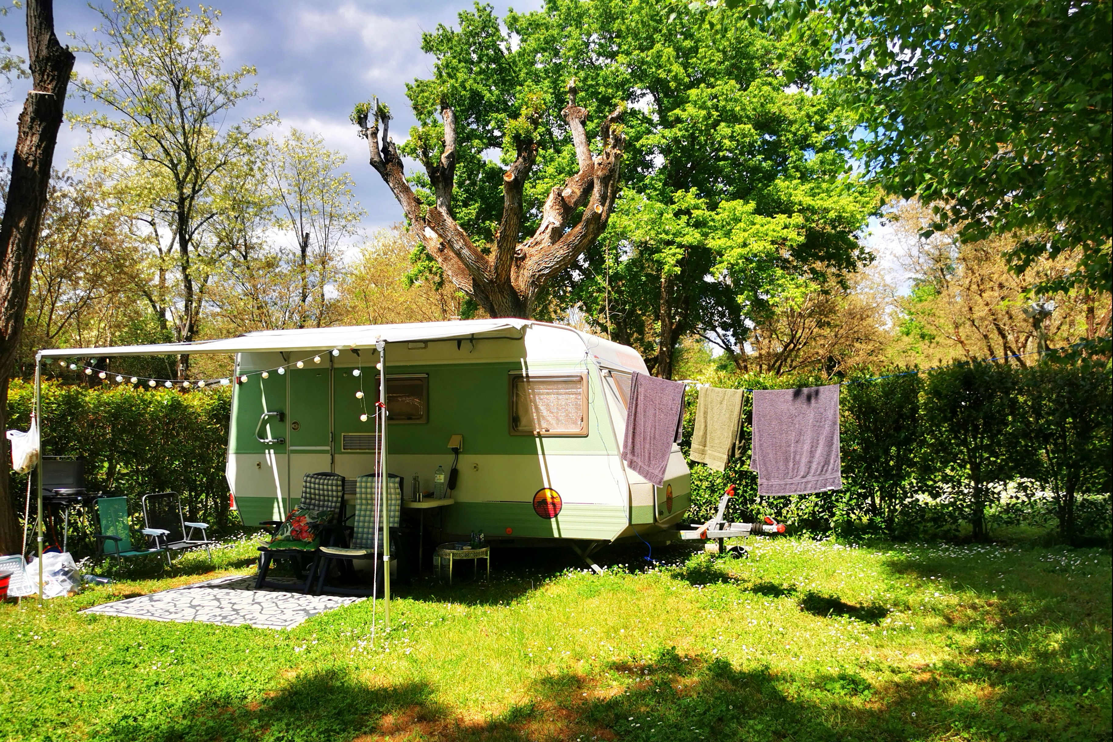 Wohnwagen mit Vorzelt auf einem Campingplatz, Rasenfläche und Sitzbank, umgeben von Bäumen und Hecken im Garten.