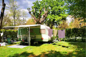 Wohnwagen mit Vorzelt auf einem Campingplatz, Rasenfläche und Sitzbank, umgeben von Bäumen und Hecken im Garten.