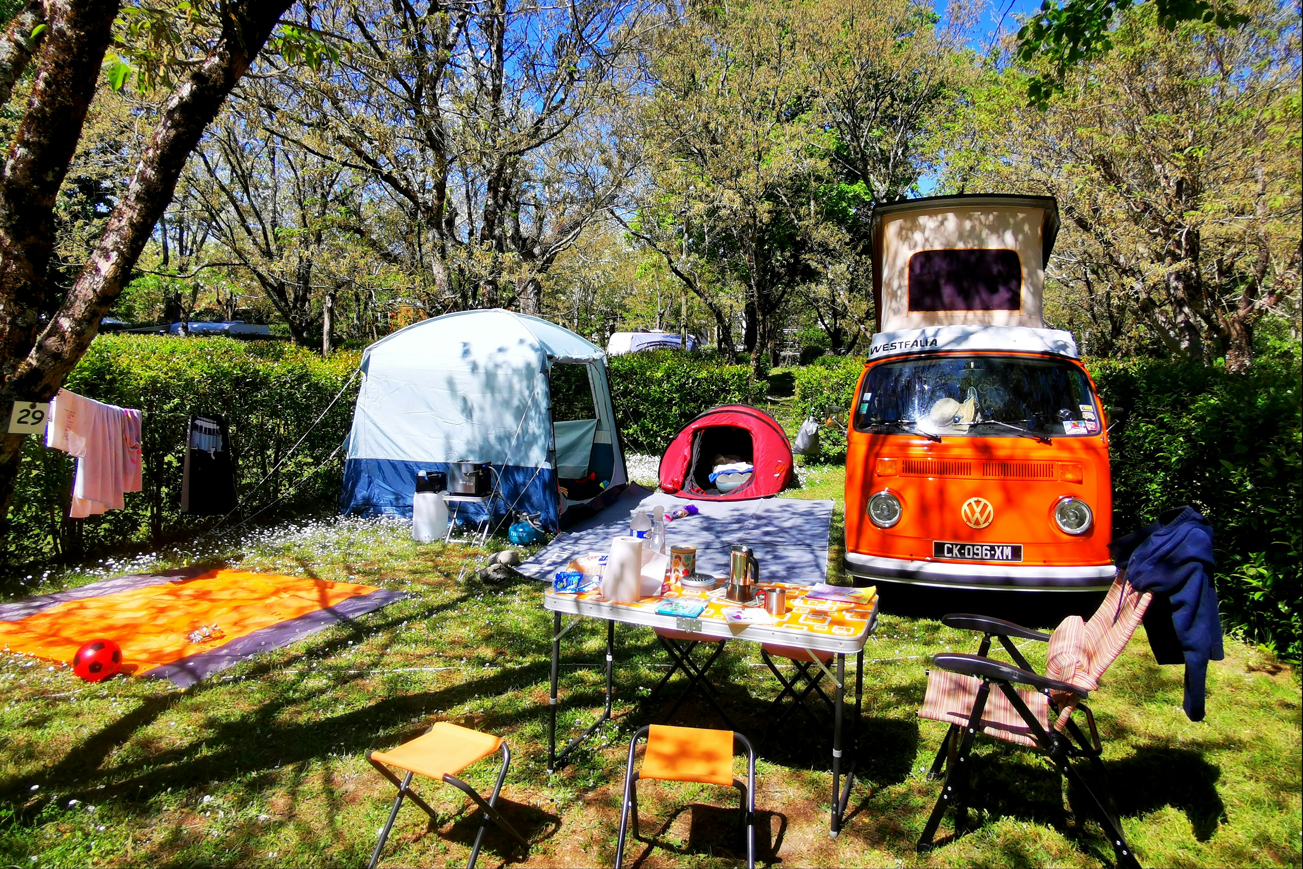 Campingplatz mit weißem Zelt, buntem Tisch und Stühlen sowie einem roten und einem orangefarbenen VW-Bus zwischen Bäumen.