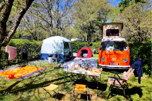 Campingplatz mit weißem Zelt, buntem Tisch und Stühlen sowie einem roten und einem orangefarbenen VW-Bus zwischen Bäumen.