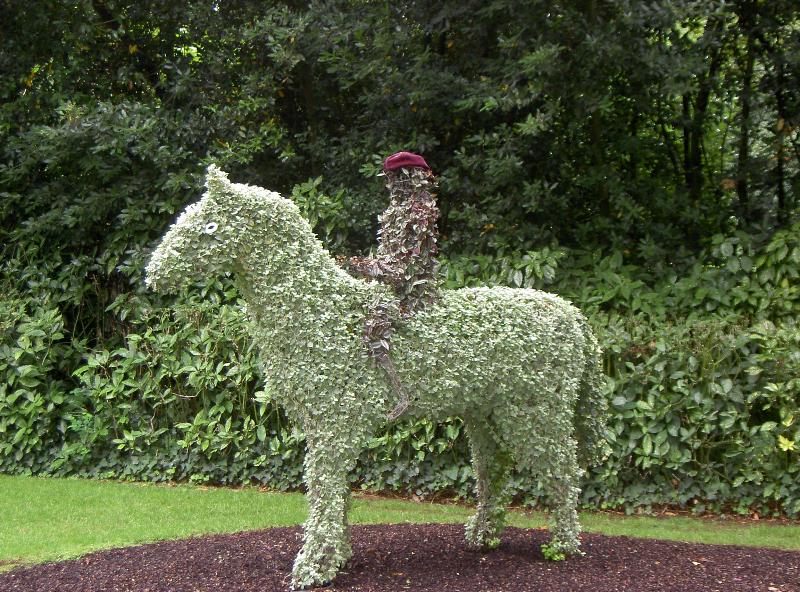 Grüne Tier-Skulptur aus Sträuchern im Garten, mit Blumen und Hecken im Hintergrund.