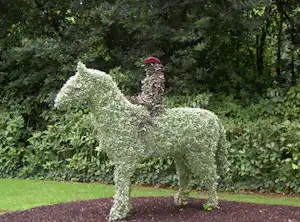 Grüne Tier-Skulptur aus Sträuchern im Garten, mit Blumen und Hecken im Hintergrund.