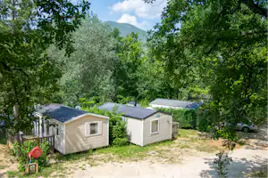 Zwei Bungalows auf einem Campingplatz im Grünen, mit Kiesweg, Bäumen und Blick auf Berge in der Ferne.