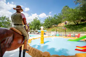 Spielzeugpferd am Camping-Pool, im Hintergrund Kinderbereich mit bunten Wasserspielzeugen und Bäumen.