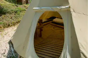 Offenes Tipi-Zelt mit Holzliege und Vorhang, im Hintergrund ein weiteres Zelt, umgeben von Bäumen und Kiesweg.