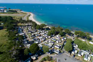 Campinganlage am Meer mit Reihen von Ferienhäusern, schmalem Strand und kurviger Straße entlang der Küste.