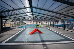 Überdachter Pool mit roter Rutsche, Liegen und Pflanzenkübeln unter einem Glasdach im Campingbereich.