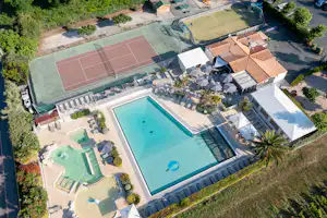 Luftaufnahme eines Campingplatzes mit großem Pool, Sportplätzen, Liegen und mehreren Gebäuden am Rand von Grünflächen.