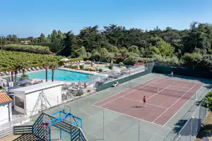 Großer Tennisplatz und Poolbereich auf dem Campingplatz, mit Liegen, Umzäunung und Blick auf Bäume und Hecken.