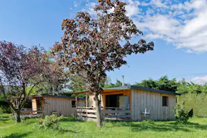 Holzbungalow im Grünen mit Terrasse, großen Fenstern und Grillkamin, umgeben von Bäumen und Rasen.