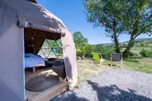 Zelt mit Holzliege und Vorhang, daneben Campingstühle auf Kiesfläche, im Hintergrund Wiese und Bäume.