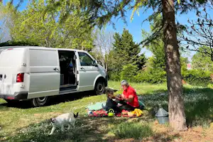 Weißer Camping-Van am Stellplatz im Grünen, daneben Decke und Spielzeug, eine Person sitzt mit Hund im Gras.