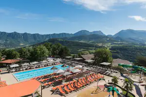 Großes Camping-Schwimmbecken mit vielen Liegen, Sonnenschirmen und Blick auf grüne Berge in der Ferne.