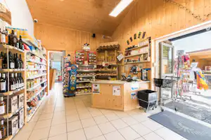 Holzverkleideter Verkaufsraum mit Regalen voller Lebensmittel und Getränken, Theke und Kasse, daneben ein Grill im Eingangsbereich.