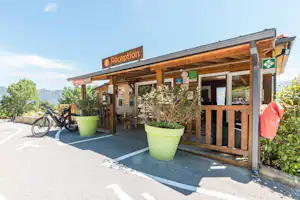 Rezeption eines Campingplatzes mit Holzveranda, Sitzbereich und großen Pflanzkübeln, daneben ein Fahrrad und Berge im Hintergrund.