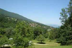 Blick ins Tal: grüner Wald, ein Dorf mit Häusern und roten Dächern, im Hintergrund Berge und Wasserfläche.