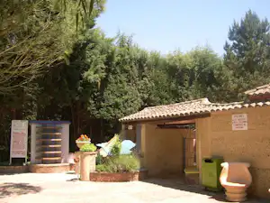 Haus mit überdachter Terrasse am Campingplatz, daneben Töpfe und Wege, umgeben von Bäumen und Sträuchern.