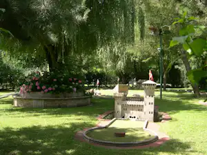 Minigolfanlage im grünen Park mit kleinen Burgen aus Kunststoff, Rasenfläche, Blumenbeeten und Bäumen.