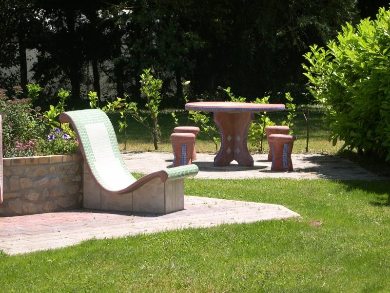 Sitzgruppe auf einer gepflasterten Terrasse im Campinggarten: weiße Liege, Tisch und Hocker, umgeben von Büschen und Rasen.