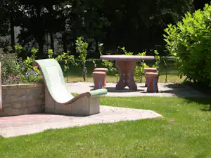 Sitzgruppe auf einer gepflasterten Terrasse im Campinggarten: weiße Liege, Tisch und Hocker, umgeben von Büschen und Rasen.