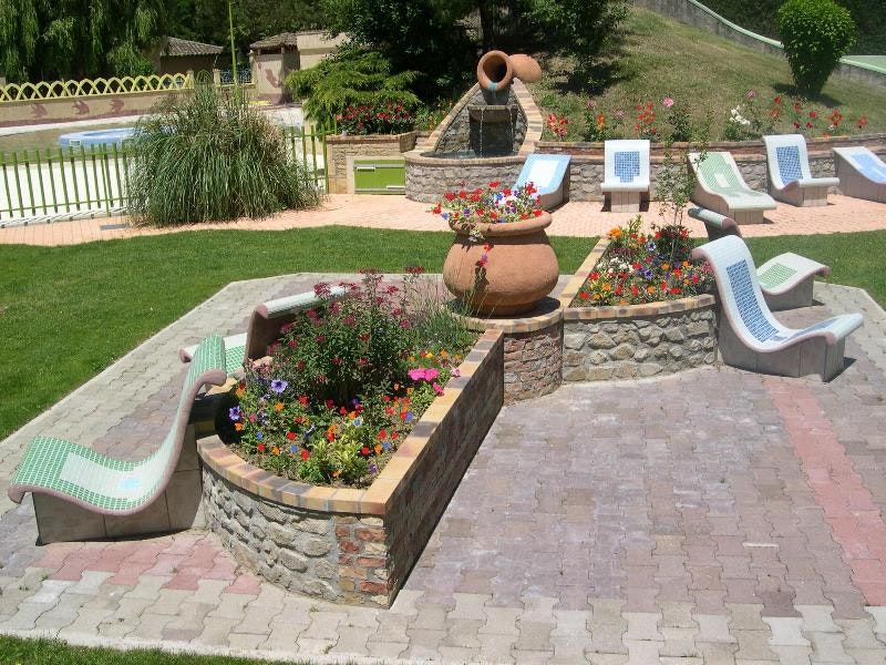Campinggarten mit Steinterrasse, Brunnen und Blumenbeeten; daneben mehrere Liegestühle und ein Poolbereich hinter dem Zaun.