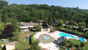 Luftaufnahme einer Campinganlage mit Pool, mehreren Gebäuden, Liegewiese und Wegen im Grünen, umgeben von Bäumen und Hügeln.