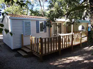 Wohnwagen mit Veranda und Holzgeländer, seitlich ein Fenster und eine Tür, umgeben von Bäumen im Campingbereich.