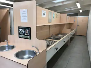 Küchenzeile im Campingbereich mit zwei Spülbecken, Grillplatte und Schränken, daneben Fliesenboden und Schilder an der Wand.