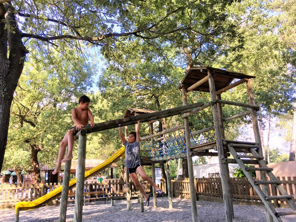 Holz-Spielgerüst auf einem Campingplatz im Grünen, Kinder klettern, im Hintergrund Bäume und ein gelbes Spielseil.