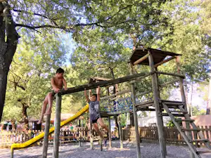 Holz-Spielgerüst auf einem Campingplatz im Grünen, Kinder klettern, im Hintergrund Bäume und ein gelbes Spielseil.