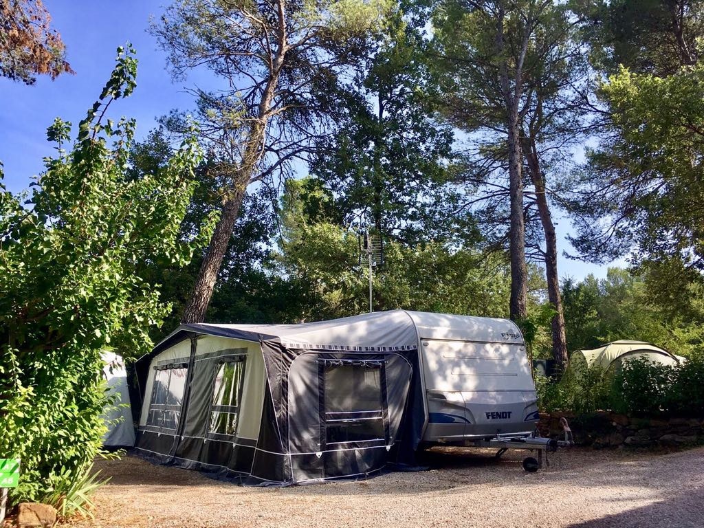Wohnwagen mit Vorzelt auf einem Campingplatz-Stellplatz, umgeben von Bäumen und Sträuchern, mit Schotterfläche.