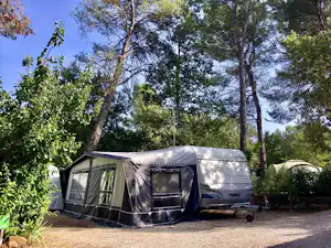 Wohnwagen mit Vorzelt auf einem Campingplatz-Stellplatz, umgeben von Bäumen und Sträuchern, mit Schotterfläche.