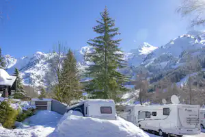 Wohnmobile im Schnee auf dem Campingplatz, im Hintergrund verschneite Berge und ein hoher Nadelbaum.