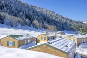 Winterlicher Campingplatz mit mehreren Bungalows aus Holz, verschneiten Wegen und Bergen im Hintergrund.