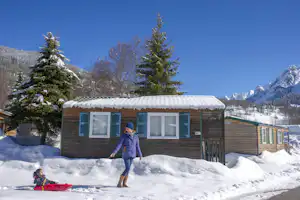 Winterliches Campinghaus mit weißem Dach und großen Fenstern, davor Schnee und ein Schlitten im Vordergrund.