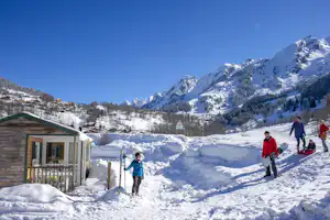 Winterlicher Campingplatz mit kleinem Haus, verschneitem Weg und Bergen im Hintergrund, mehrere Personen laufen durch den Schnee.