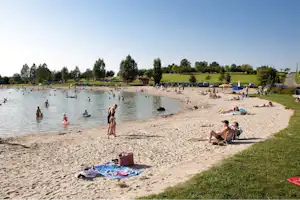 Badestrand am See mit vielen Badegästen, Liegen und Sonnenschirmen, daneben Rasen und Zelte am Campingplatz.