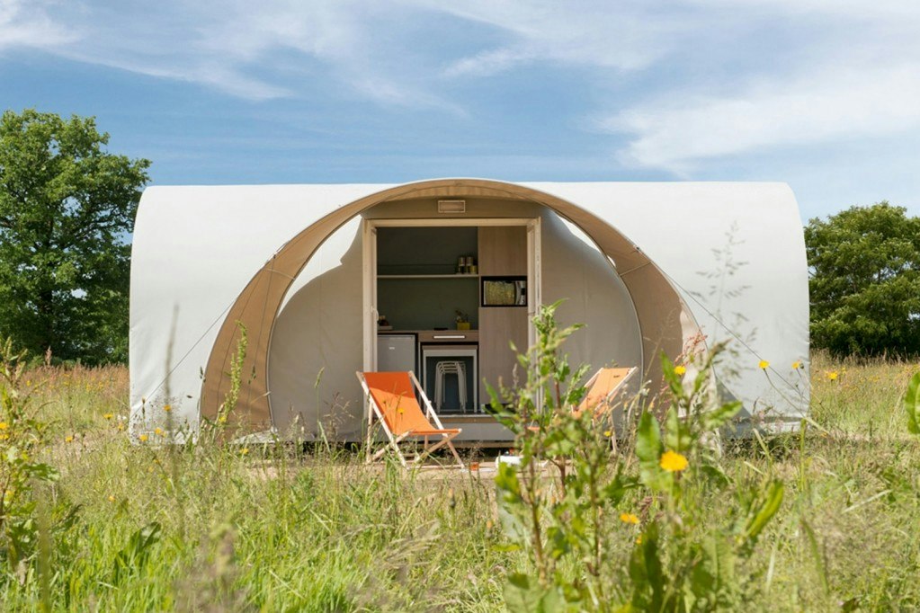 Offenes Campingzelt mit Blick ins Innere, orange Liegestühle und Küchenzeile, umgeben von Wiese und Blumen.