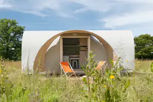 Offenes Campingzelt mit Blick ins Innere, orange Liegestühle und Küchenzeile, umgeben von Wiese und Blumen.