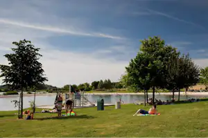 Camping am See: Wiese mit Bäumen, Spielbereich und Menschen am Ufer, im Hintergrund Wasser und Liegewiese.
