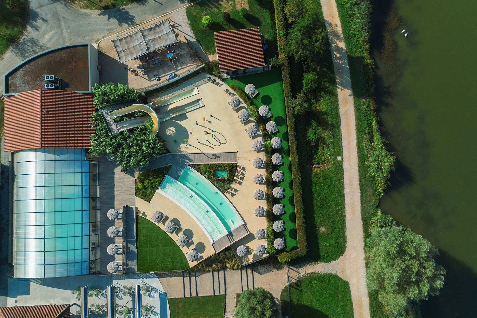 Luftaufnahme eines Campingplatzes mit Schwimmbecken, Sonnendeck, Liegen und Beckenbereich neben einem großen Gebäude und Wegen.