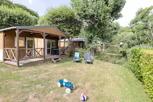 Holz-Bungalow mit überdachter Terrasse, zwei Stühlen und Tisch im Garten, umgeben von Bäumen und Grün.